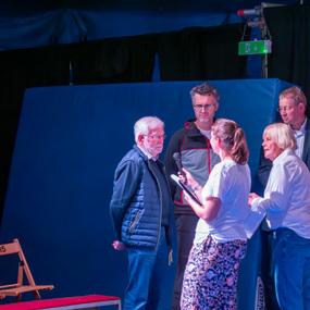 Vier Personen stehen auf einer Bühne, während eine Frau mit einem Mikrofon in der Hand spricht.