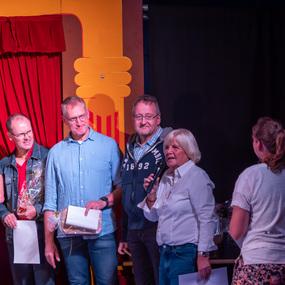 Fünf Personen stehen auf einer Bühne, während eine Frau mit Mikrofon spricht. Rote Vorhänge und eine dunkle Kulisse sind sichtbar.