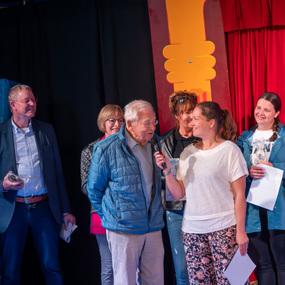 Eine Gruppe von sechs Personen steht auf einer Bühne, während eine Frau mit Mikrofon einen älteren Mann interviewt.