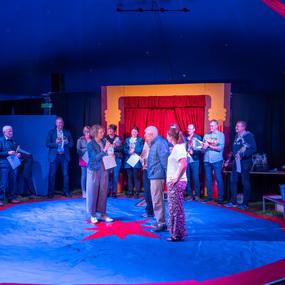 Menschen stehen auf einer blauen Zirkusfläche, applaudieren und halten Dokumente, während eine Person spricht.