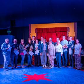 Gruppe von Personen mit Urkunden und Trophäen auf einer Bühne in einem Zirkuszelt vor rotem Vorhang.