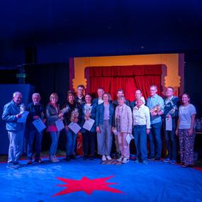 Gruppe von Personen steht auf einer blauen Zirkusmatte vor rotem Vorhang, hält Urkunden und Preise in der Hand.