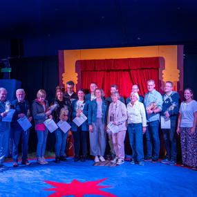 Gruppenfoto von 15 Personen auf einer Bühne, alle halten Urkunden und Blumen, vor einem roten Vorhang.