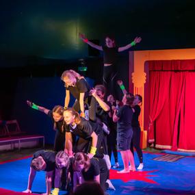 Gruppe von Tänzern in Schwarz bildet einen menschlichen Turm auf einer bunten Zirkusmatte vor einem roten Vorhang.