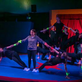 Acht junge Erwachsene in Schwarz führen akrobatische Übungen auf einer blauen Matte unter rot beleuchtetem Vorhang aus.