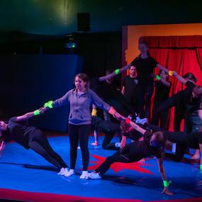 Acht Akrobaten in Schwarz führen eine Gruppenübung auf einer blauen Matte unter bunten Lichtern durch.