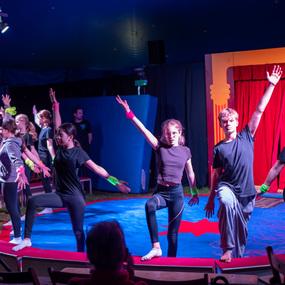 Sechs junge Darstellerinnen und Darsteller in Schwarz mit bunten Arm-Accessoires stehen beim Auftritt in einem Zirkuszelt.