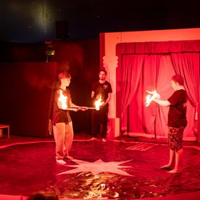 Drei Akrobaten im Zirkus führen eine Feuerjonglage unter rotem Licht auf einer Bühne vor.
