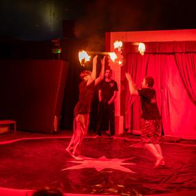 Zwei Künstler jonglieren mit brennenden Fackeln auf einer roten Zirkusfläche, ein Zuschauer beobachtet im Hintergrund.