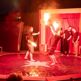 Zwei Artisten führen im Zirkus eine Feuershow mit brennenden Fackeln auf einer roten Bühne vor mehreren Zuschauern auf.
