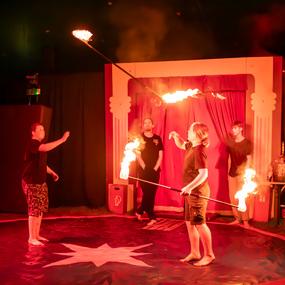 Zwei Kinder jonglieren mit brennenden Fackeln auf einer Zirkusfläche, während Zuschauer im Hintergrund beobachten.