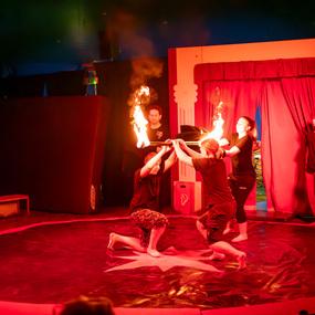 Zwei Künstler führen eine Feuerakrobatik-Nummer in einem Zirkuszelt mit rotem Licht auf. Zuschauer stehen im Hintergrund.