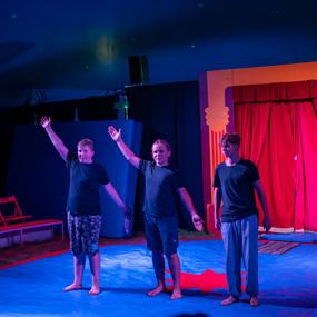 Drei jugendliche Darsteller stehen auf einer blauen Bühne und heben ihre Arme in einem Zirkuszelt mit rotem Vorhang.