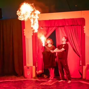 Zwei Künstler in schwarzen T-Shirts führen eine Feuerspuck-Performance vor rotem Bühnenvorhang durch.