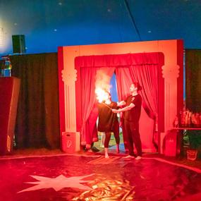 Darsteller entfachen Flammen während einer Zirkusaufführung vor rotem Vorhang und einer beleuchteten Kulisse.