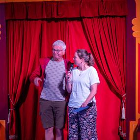 Zwei Personen stehen auf einer Bühne vor einem roten Vorhang, die Frau hält ein Mikrofon und schaut den Mann an.