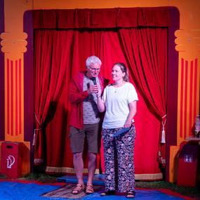 Zwei Personen stehen auf einer Bühne vor roten Vorhängen und sprechen ins Mikrofon, umgeben von Bühnenrequisiten.