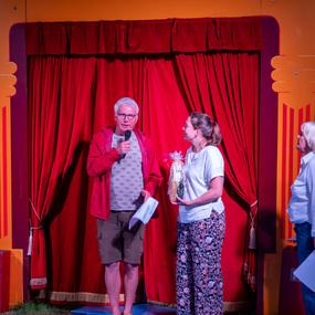 Drei Personen stehen vor einem roten Vorhang. Ein Mann spricht ins Mikrofon, während eine Frau ein Geschenk hält.