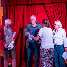 Vier Personen stehen auf einer Bühne vor roten Vorhängen; zwei erhalten Preise, während eine Frau mit Mikrofon spricht.