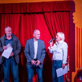Vier Personen stehen auf einer Bühne vor einem roten Vorhang, eine hält ein Mikrofon und spricht.