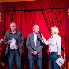 Vier Personen stehen auf einer Bühne vor rotem Vorhang, während eine Frau mit Mikrofon einen Mann befragt.