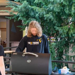 Ein junger Mann mit lockigem Haar steht vor einem Grill, während Jugendliche im Hintergrund beobachtend schauen.