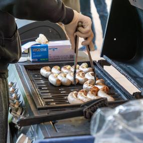 Grill mit verschiedenen gegrillten Würstchen, während eine Hand mit Zange sie wendet. Im Hintergrund sind andere Personen sichtbar.