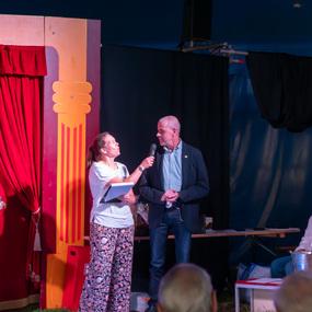 Eine Frau spricht mit einem Mikrofon, während zwei Männer und eine Frau auf einer Bühne sitzen. Roter Vorhang im Hintergrund.