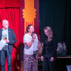 Vier Personen stehen auf einer Bühne, während eine Frau mit Mikrofon spricht. Rote Vorhänge im Hintergrund.