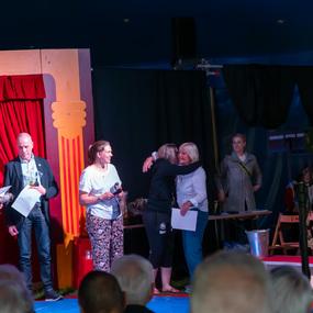 Mehrere Personen stehen auf einer Bühne in einem Zirkuszelt, einige umarmen sich und halten Urkunden in den Händen.