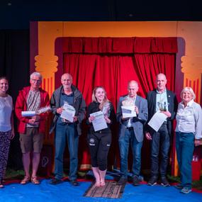 Gruppe von sieben Personen steht vor rotem Vorhang auf Bühne, alle halten Urkunden oder Preise in den Händen.