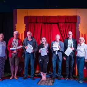 Sieben Personen stehen vor einem roten Vorhang, halten Urkunden und Preise in der Hand, auf einer bunten Bühne.