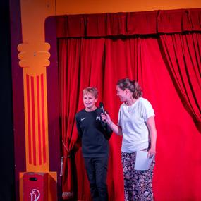 Kind und Erwachsene stehen vor rotem Vorhang, die Frau hält ein Mikrofon und spricht mit dem Jungen.