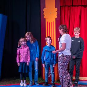 Vier Kinder stehen auf einer Bühne, während eine Erwachsene mit Mikrofon spricht. Rote Vorhänge im Hintergrund.