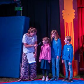 Eine Frau interviewt vier Kinder auf einer bunten Zirkusbühne mit einem blauen Hintergrund.
