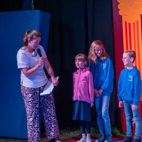 Eine Moderatorin steht auf einer Bühne und spricht zu drei Kindern in blauen und rosa Hoodies.