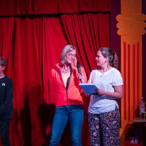 Drei Personen stehen auf einer Bühne vor einem roten Vorhang; eine Frau spricht ins Mikrofon, während der Junge neben ihr steht.