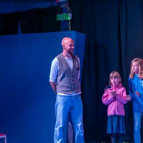 Vier Kinder und ein Mann stehen auf einer Bühne mit buntem Hintergrund, während sie miteinander interagieren.