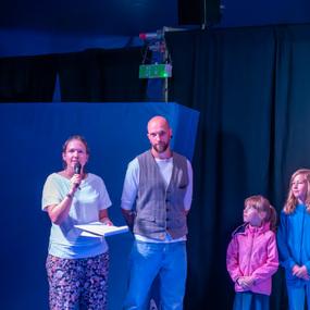 Frau mit Mikrofon spricht auf einer Bühne, neben ihr stehen ein Mann und zwei Mädchen in bunter Kleidung.