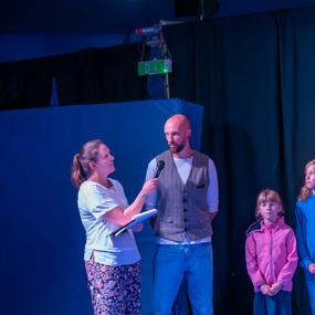 Frau interviewt einen Mann im Zirkuszelt, während zwei Kinder aufmerksam danebenstehen.