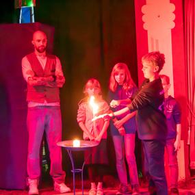 Kind mit brennendem Feuerstäbchen auf einer Bühne umgeben von mehreren Zuschauer:innen in einer bunten Beleuchtung.