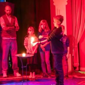 Junge zeigt eine kleine Flamme auf seiner Hand, während fünf Kinder und ein Erwachsener zuschauen. Bühne in rotem Licht.