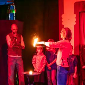 Eine Frau zeigt eine Fackel mit Flamme, während Zuschauer, darunter Kinder, gespannt beobachten. Rotes Licht im Hintergrund.