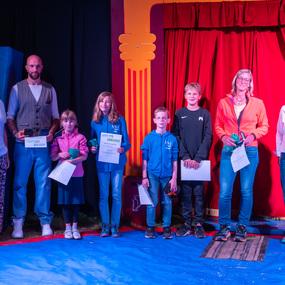 Gruppe von sieben Personen, darunter Kinder und Erwachsene, präsentiert Urkunden auf einer Bühne mit rotem Vorhang.