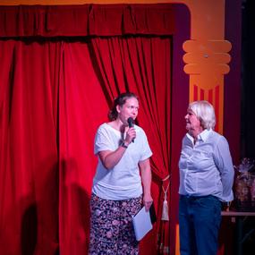 Zwei Frauen stehen vor einem roten Vorhang; eine hält ein Mikrofon, während die andere lächelt.