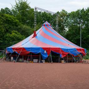 Bunt gestreifter Zirkuswagen auf einem Sportplatz, umgeben von Bäumen und mit verschiedenen Kisten vor dem Eingang.