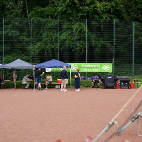 Menschen unter Pavillons sitzen und stehen auf einem sandigen Platz, im Hintergrund eine Begrünung und ein Schild.
