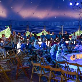 Zuschauer sitzen auf Holzstühlen im Zelt, umgeben von bunten Lichtstrahlen und blauer Zeltplane.