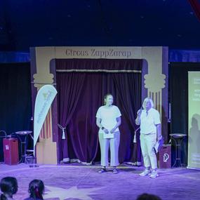 Zwei Personen stehen auf der Bühne des Circus ZappZarap und sprechen vor einem Publikum von Kindern.