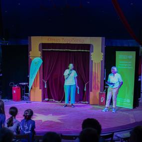 Frau mit Mikrofon spricht auf der Bühne eines Zirkus, während Zuschauer im Hintergrund sitzen.